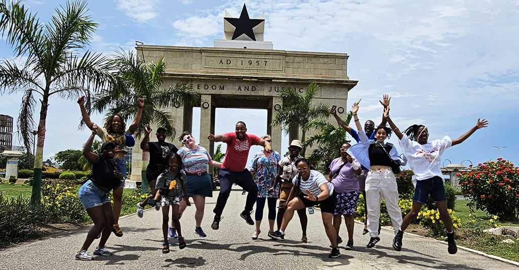 A group of 13 stand in front of the Black Star Square monument, a concrete structure that reads AD 1957. Freedom and Justice. A black star sits atop the stone structure. Green palm trees and flowers flank the monument.