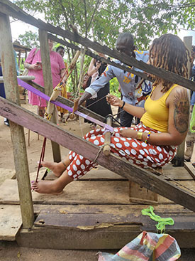 Wearing a yellow tank top and red pants with white polka dots, Nicole Corley sits at a Ghanaian weaving loom.