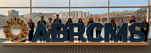 A group of student researchers stand in front of oversize letters, ABRCMS, the acronym fro the conference they are attending in Pittsburgh, Annual Biomedical Research Conference for Minoritized Scientists. Behind them are a river and cityscape