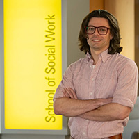 A man stands with his arms crossed in front of a gold sign that reads School of Social Work