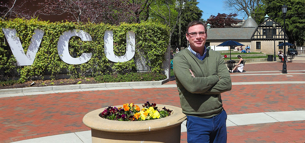 VCU interim dean Gary Cuddeback, wearing a green sweater and blue pants, stands outside by a large planter with colorful flowers, with a VCU sign in the background.