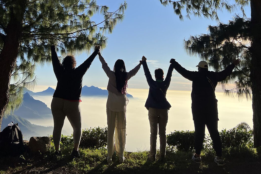 Four people clasp hands as they look out on the sun rising over cloud-filled mountains.