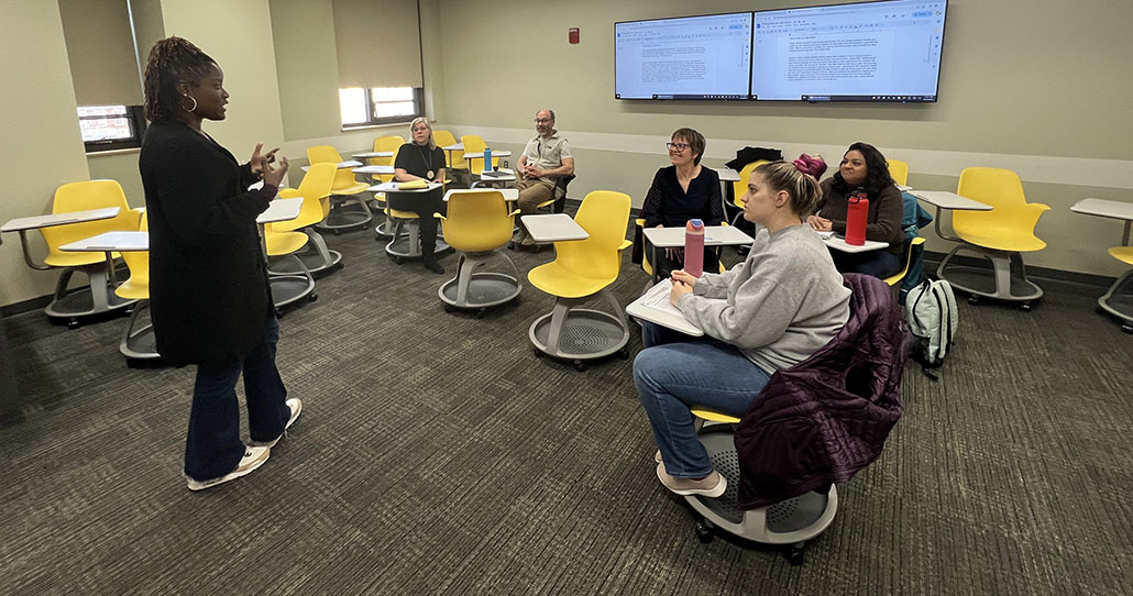 An instructor stands in front of a class talking to five students seated in gold chairs.