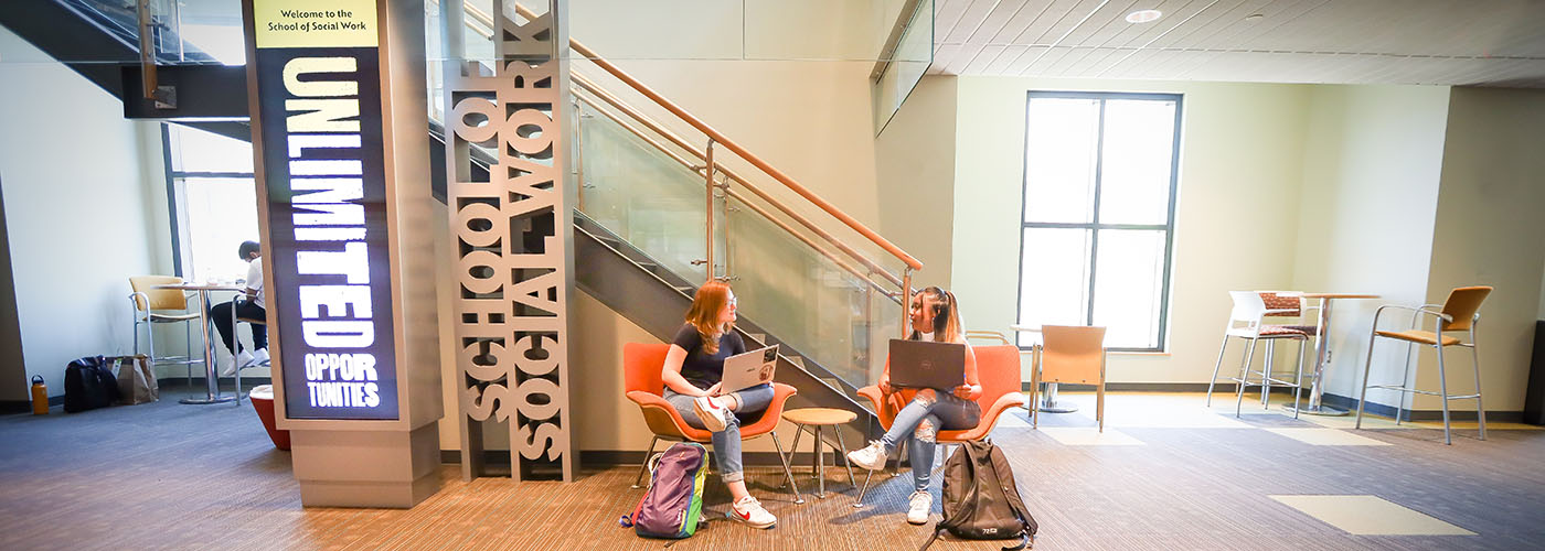 Two social work students talk while sitting on orange chairs with their laptops. To the left are a School of Social Work sign and a digital display reading, 