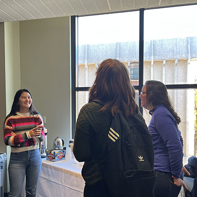 A student ambassador in a striped sweater talks with two fellow students while standing near a table with hot chocolate and coffee.