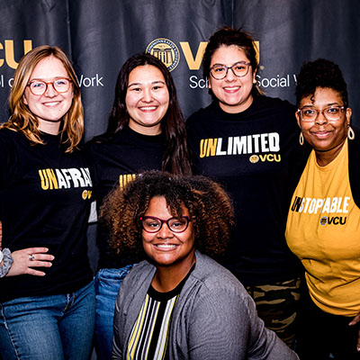 Three student ambassadors in black t-shirts stand next to the interim associate dean and the senior recruitment specialist. Two of the three student shirts read UNAFRAID and UNLIMITED. The specialist wears a gold UNSTOPPABLE shirt; the interim associate dean is wearing a black and gold striped top with a light gray sweater