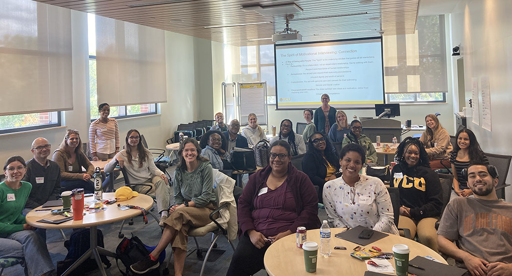 Students, faculty and staff sitting in a classroom, with a large screen showing a presentation in the back of the room.